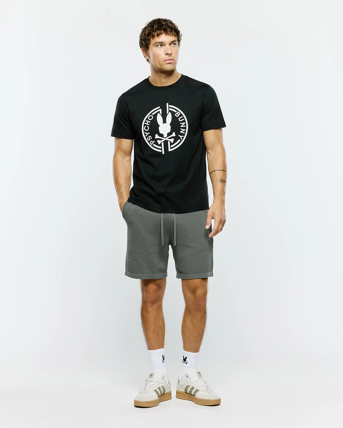 Mens Santa Fe Bunny Graphic Tee, a black classic fit premium jersey t-shirt featuring a large white circular Psycho Bunny skull logo graphic on the chest, complemented by grey textured knit drawstring shorts with a cuffed hem.