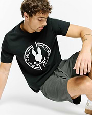 Mens Santa Fe Bunny Graphic Tee, a black classic-fit jersey t-shirt featuring a large white circular Psycho Bunny skull and crossbones graphic on the chest, styled with dark grey waffle knit drawstring shorts.