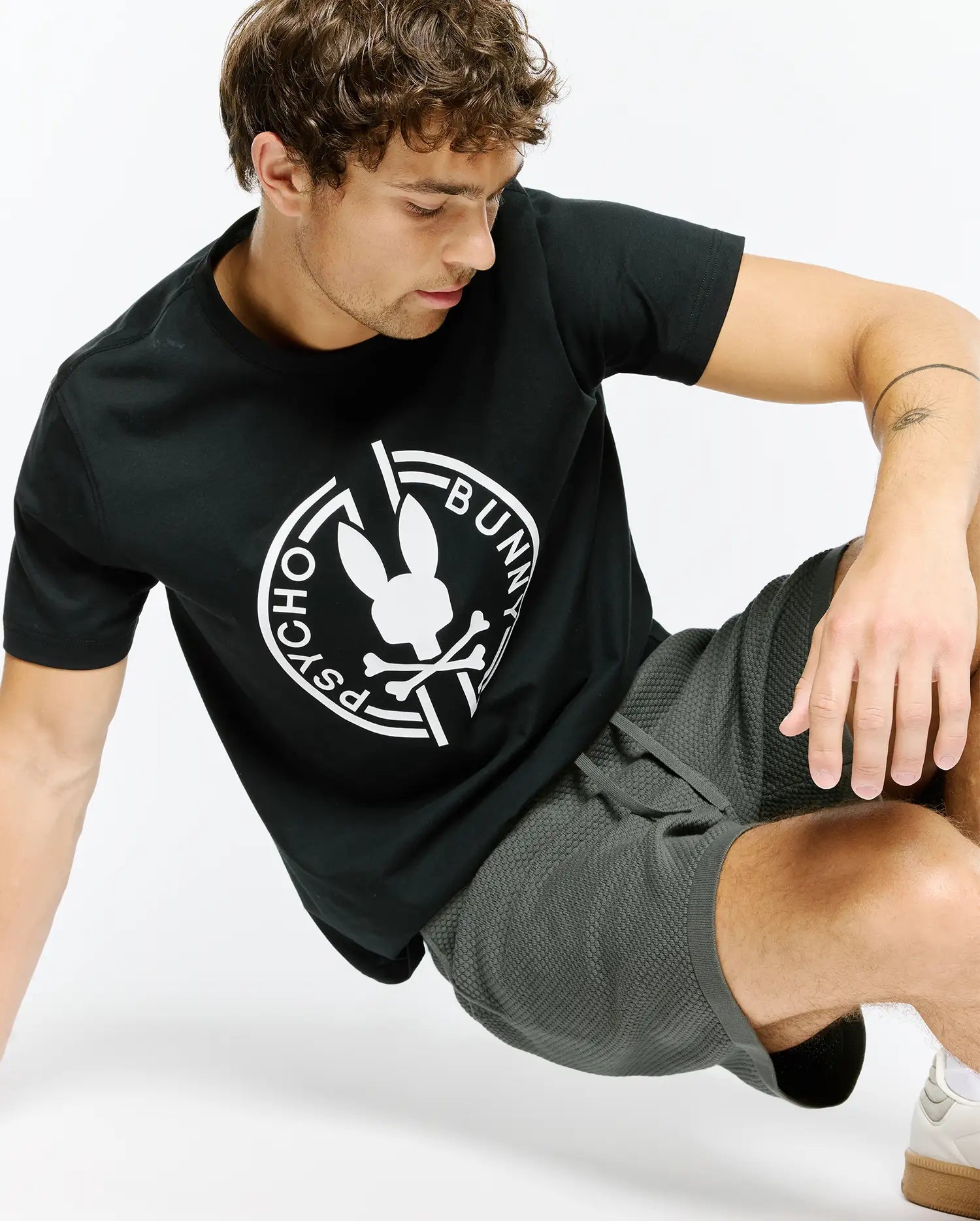 Mens Santa Fe Bunny Graphic Tee, a black classic-fit jersey t-shirt featuring a large white circular Psycho Bunny skull and crossbones graphic on the chest, styled with dark grey waffle knit drawstring shorts.