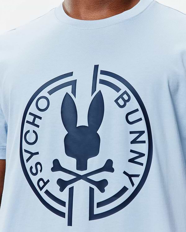 Mens Santa Fe Bunny Graphic Tee, a light blue jersey knit t-shirt with a classic fit, featuring a large navy Psycho Bunny skull and crossbones logo graphic on the central chest, encircled by PSYCHO BUNNY text. (3)