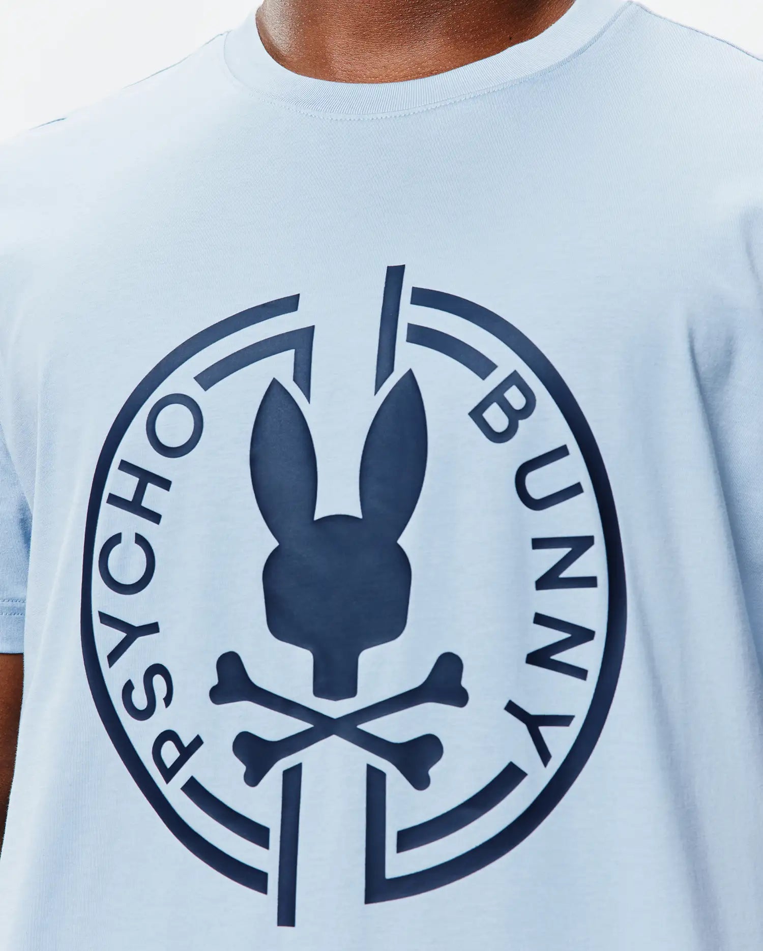 Mens Santa Fe Bunny Graphic Tee, a light blue jersey knit t-shirt with a classic fit, featuring a large navy Psycho Bunny skull and crossbones logo graphic on the central chest, encircled by PSYCHO BUNNY text. (3)