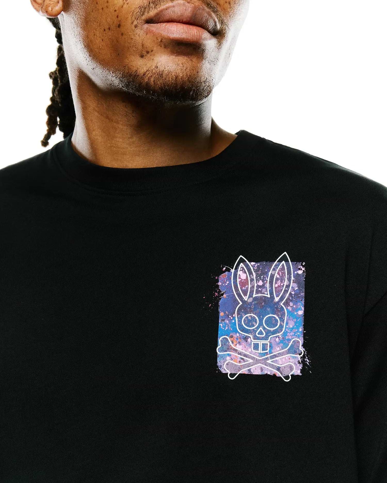 Mens Isaac Splatter Back Graphic Tee, a black jersey t-shirt with a small left chest graphic featuring a white Psycho Bunny skull and crossbones over a vibrant purple, blue, and pink splatter background.