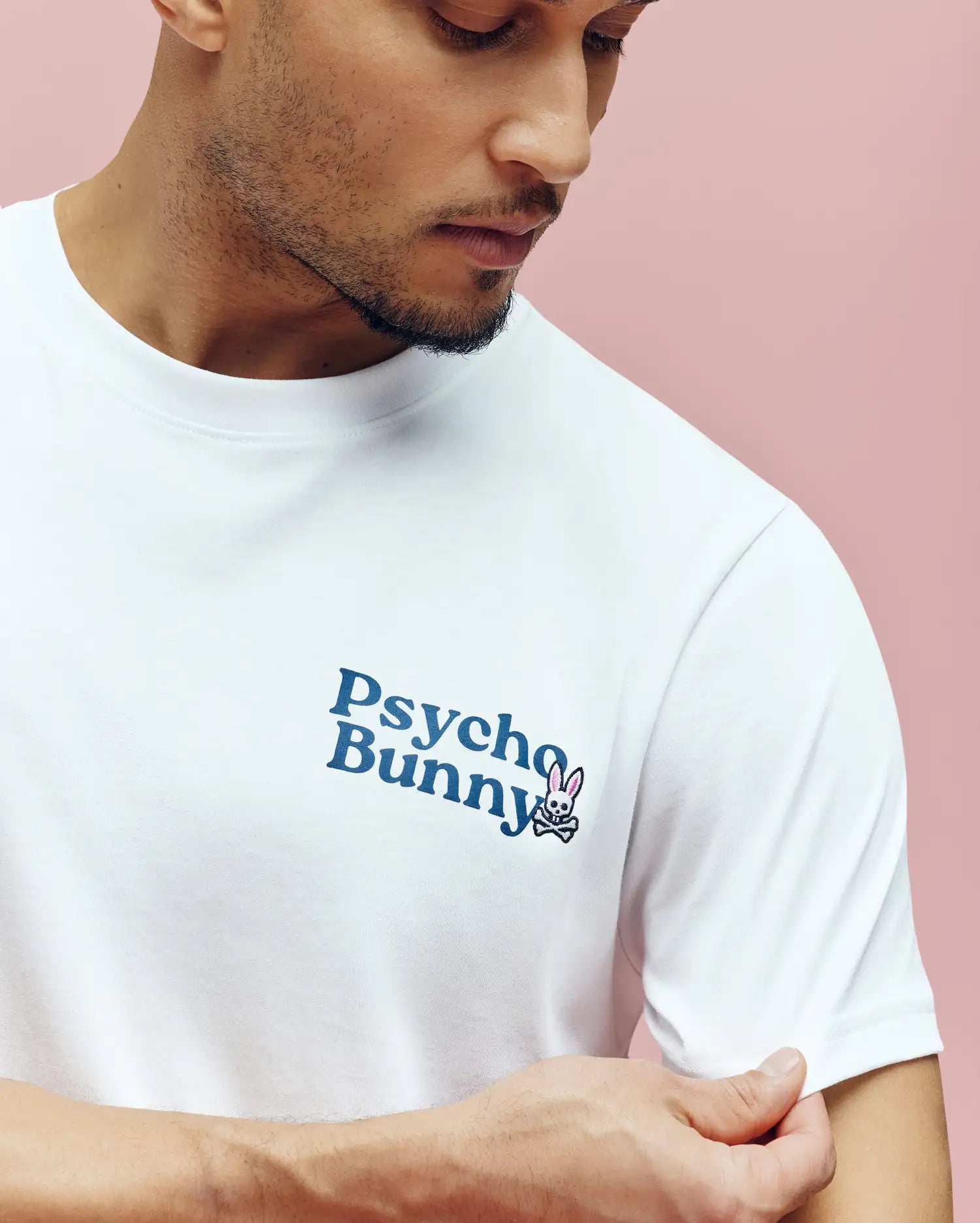 A man wearing the Psycho Bunny MENS ARCHIVE WORDMARK GRAPHIC TEE (MT0200030), a white shirt featuring the signature bunny graphic in blue and pink on the left chest, stands against a pink background.