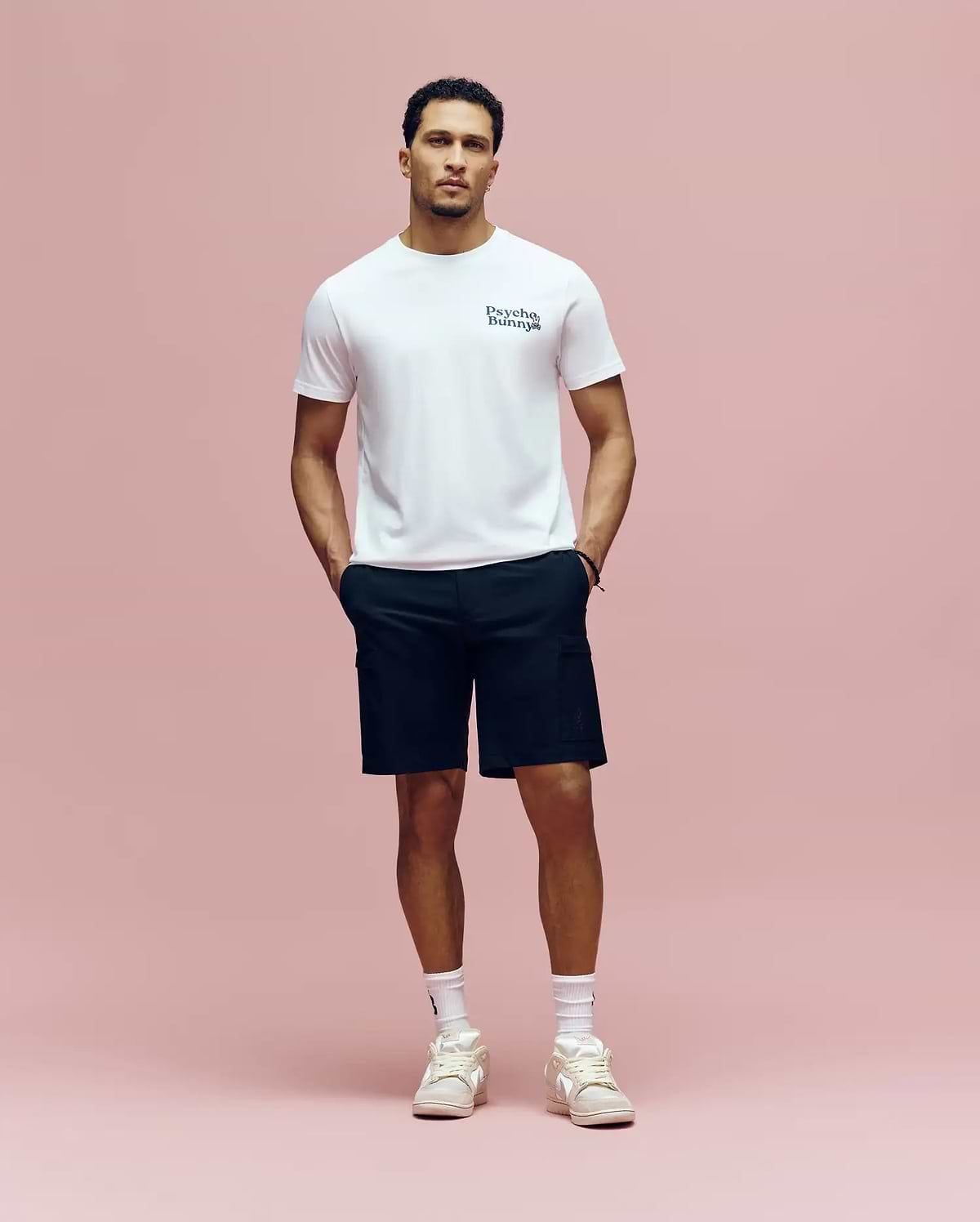 A man stands against a pink background wearing a white Psycho Bunny MENS ARCHIVE WORDMARK GRAPHIC TEE (MT0200030), black shorts, white socks, and white sneakers, with his hands in his pockets.