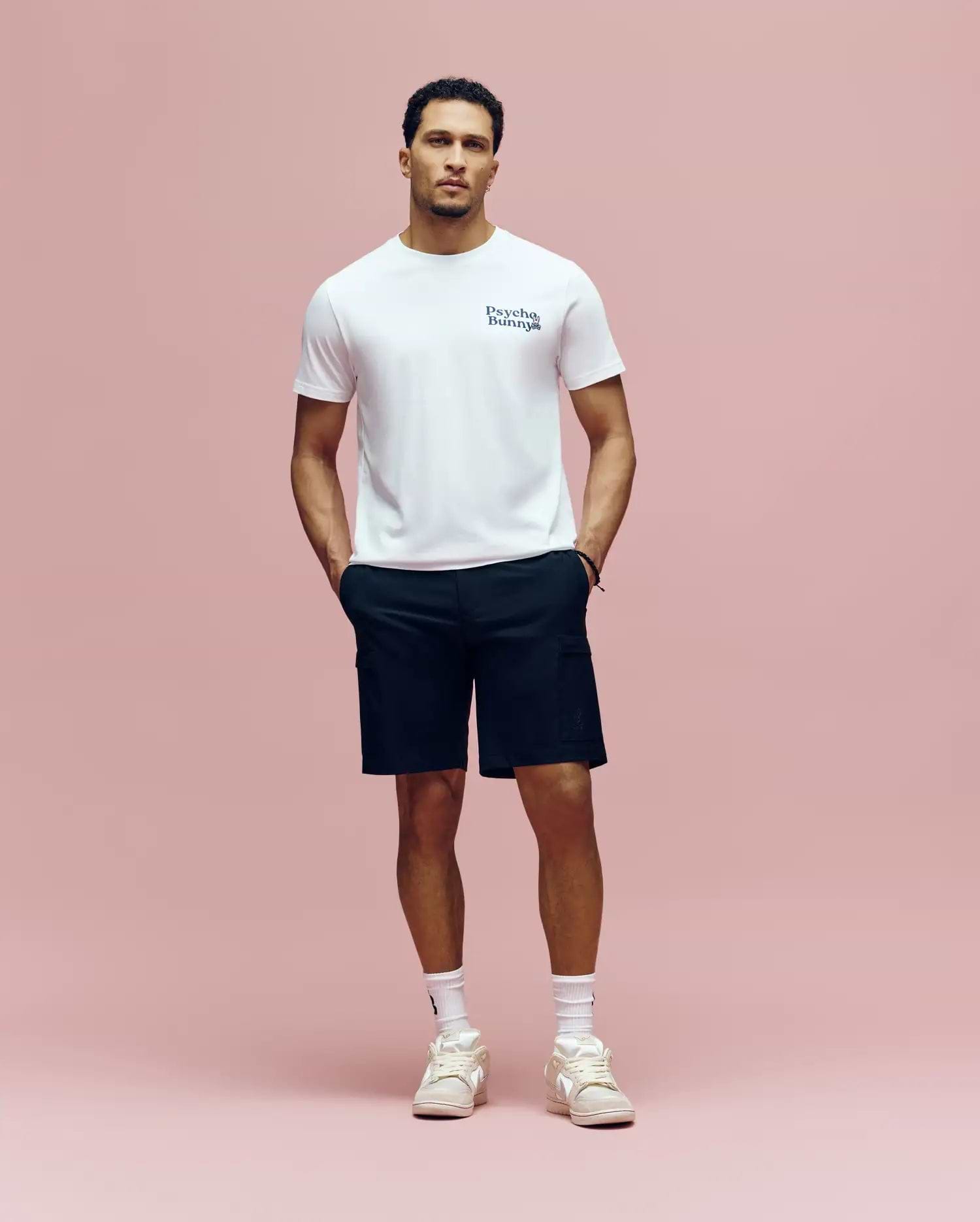 A man stands against a pink background wearing a white Psycho Bunny MENS ARCHIVE WORDMARK GRAPHIC TEE (MT0200030), black shorts, white socks, and white sneakers, with his hands in his pockets.