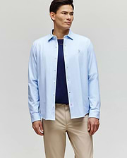 A man stands against a plain background wearing the Psycho Bunny MENS JIM JERSEY STRIPED KNIT SHIRT (MT0300041) over a navy t-shirt and beige pants, looking slightly to the side with a neutral expression.