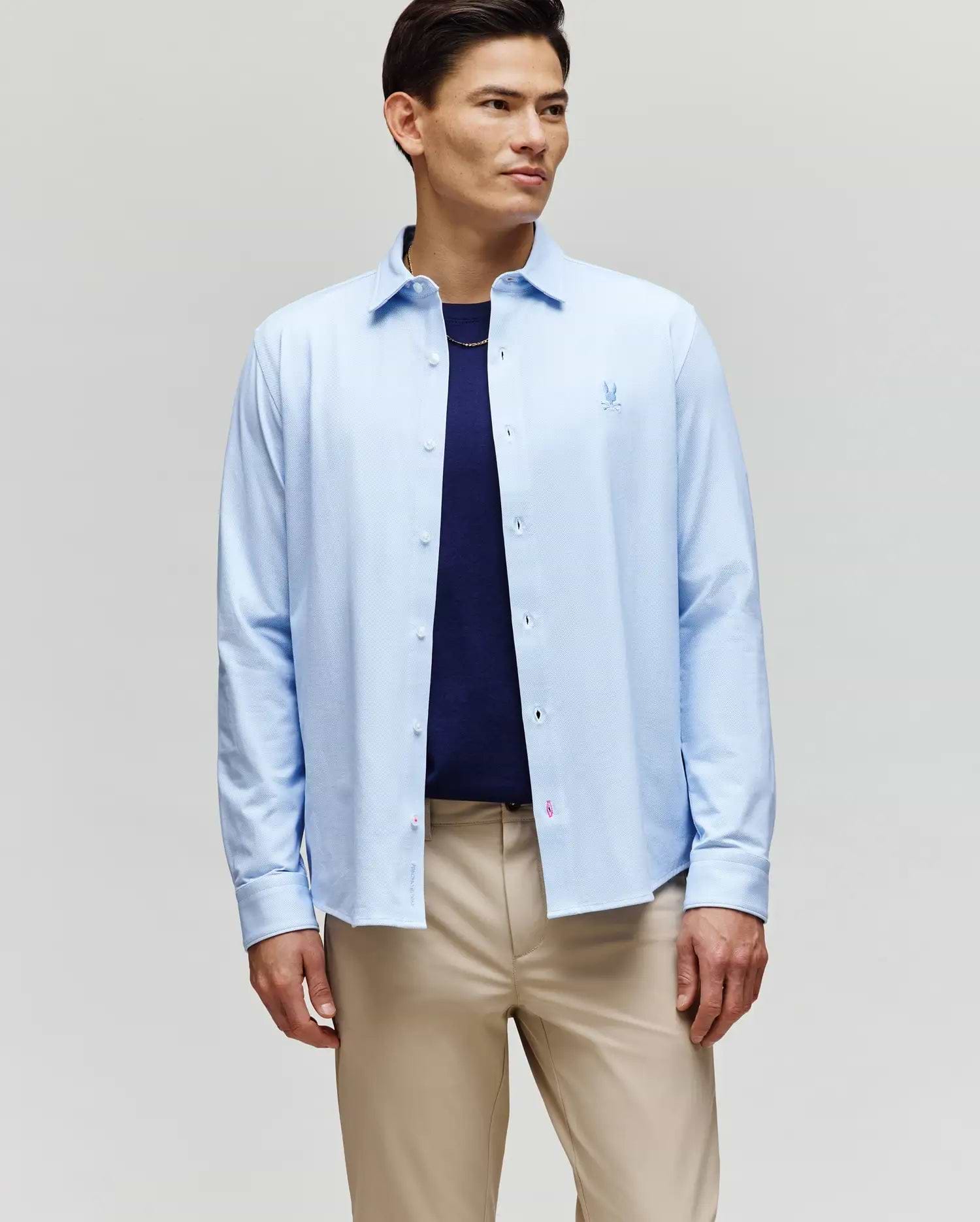 A man stands against a plain background wearing the Psycho Bunny MENS JIM JERSEY STRIPED KNIT SHIRT (MT0300041) over a navy t-shirt and beige pants, looking slightly to the side with a neutral expression.