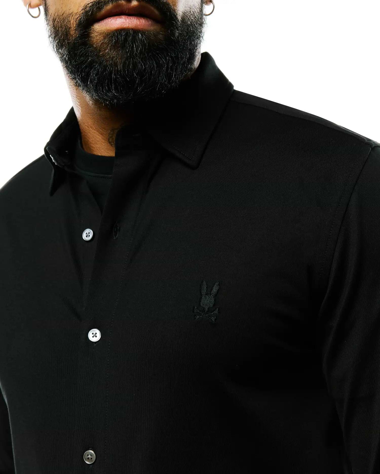 Mens Jim Jersey Knit Shirt, a black classic fit collared button-front shirt in jersey knit fabric, featuring a small tonal embroidered Psycho Bunny skull logo on the left chest and white buttons.