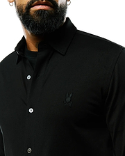 Mens Jim Jersey Knit Shirt, a black classic fit collared button-front shirt in jersey knit fabric, featuring a small tonal embroidered Psycho Bunny skull logo on the left chest and white buttons.