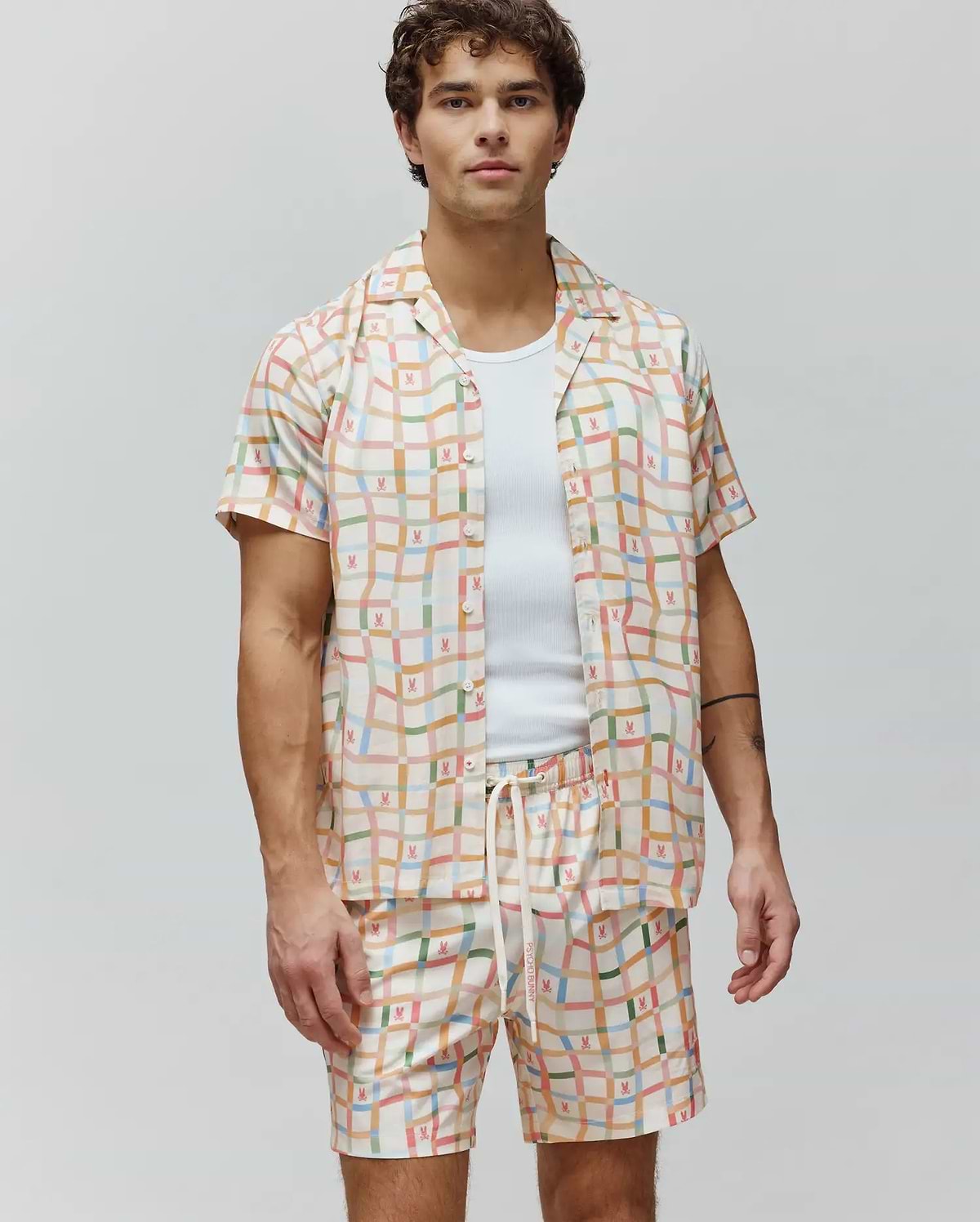 Mens Atlas Tencel Shirt, a relaxed fit cream short-sleeve button-up with a vibrant multicolor grid pattern and repeated small Psycho Bunny skull logos.