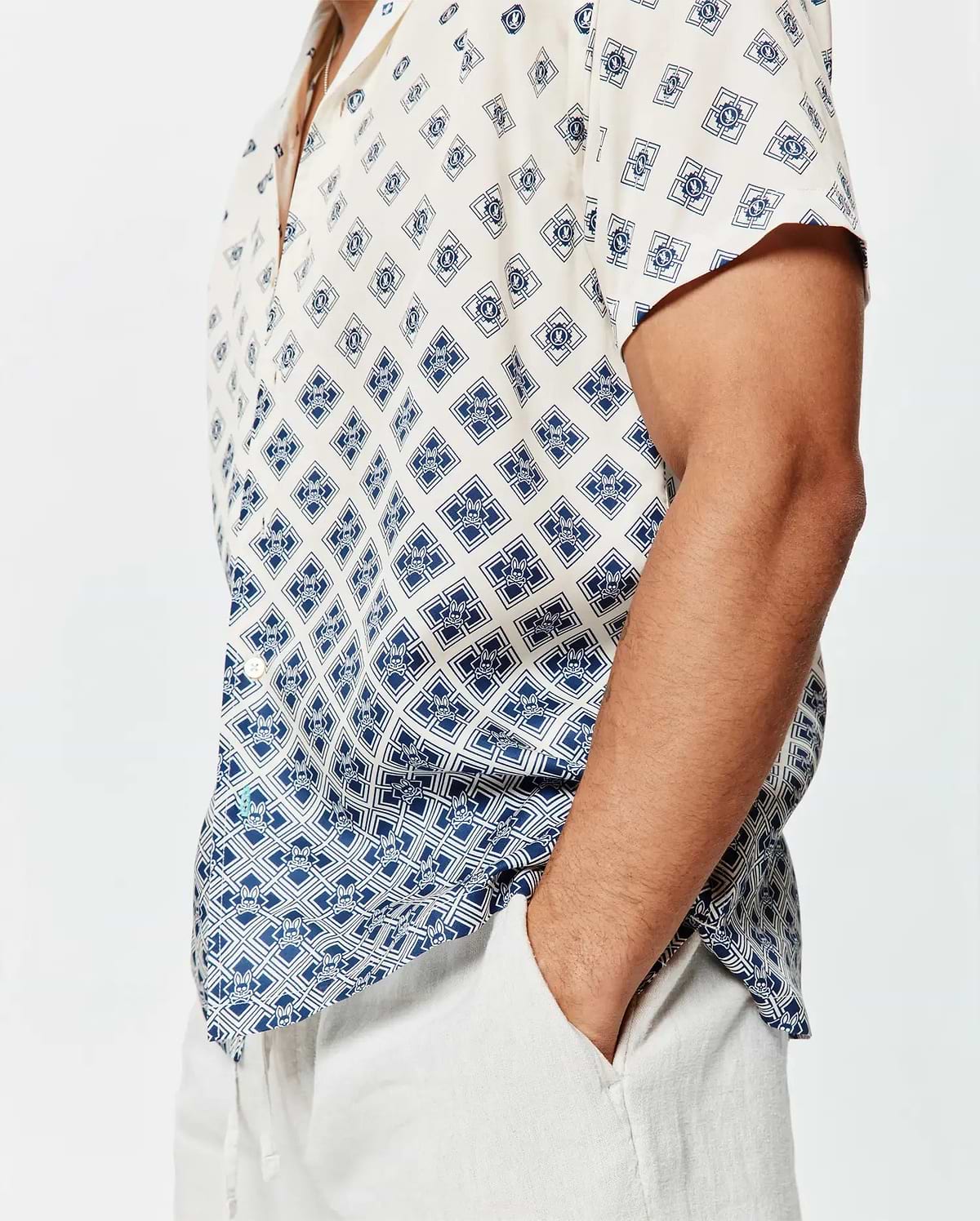 Mens Tobias Tencel Shirt, a relaxed fit cream short-sleeve button-up with a navy blue geometric and Psycho Bunny skull logo all-over print, featuring a subtle gradient effect and a small green hem tag.