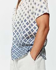 Mens Tobias Tencel Shirt, a relaxed fit cream short-sleeve button-up with a navy blue geometric and Psycho Bunny skull logo all-over print, featuring a subtle gradient effect and a small green hem tag.