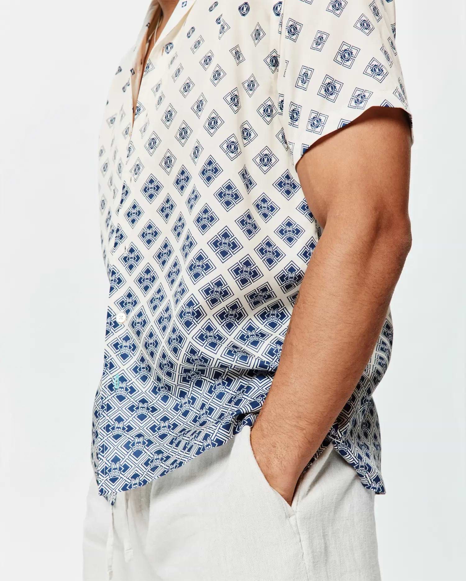 Mens Tobias Tencel Shirt, a relaxed fit cream short-sleeve button-up with a navy blue geometric and Psycho Bunny skull logo all-over print, featuring a subtle gradient effect and a small green hem tag.