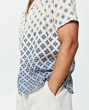 Mens Tobias Tencel Shirt, a relaxed fit cream short-sleeve button-up with a navy blue geometric and Psycho Bunny skull logo all-over print, featuring a subtle gradient effect and a small green hem tag.