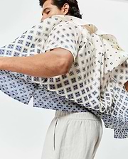 Mens Tobias Tencel Shirt, cream short-sleeve with a relaxed fit, featuring an all-over navy blue geometric pattern subtly incorporating small Psycho Bunny skull logos, transitioning to a denser pattern at the hem.