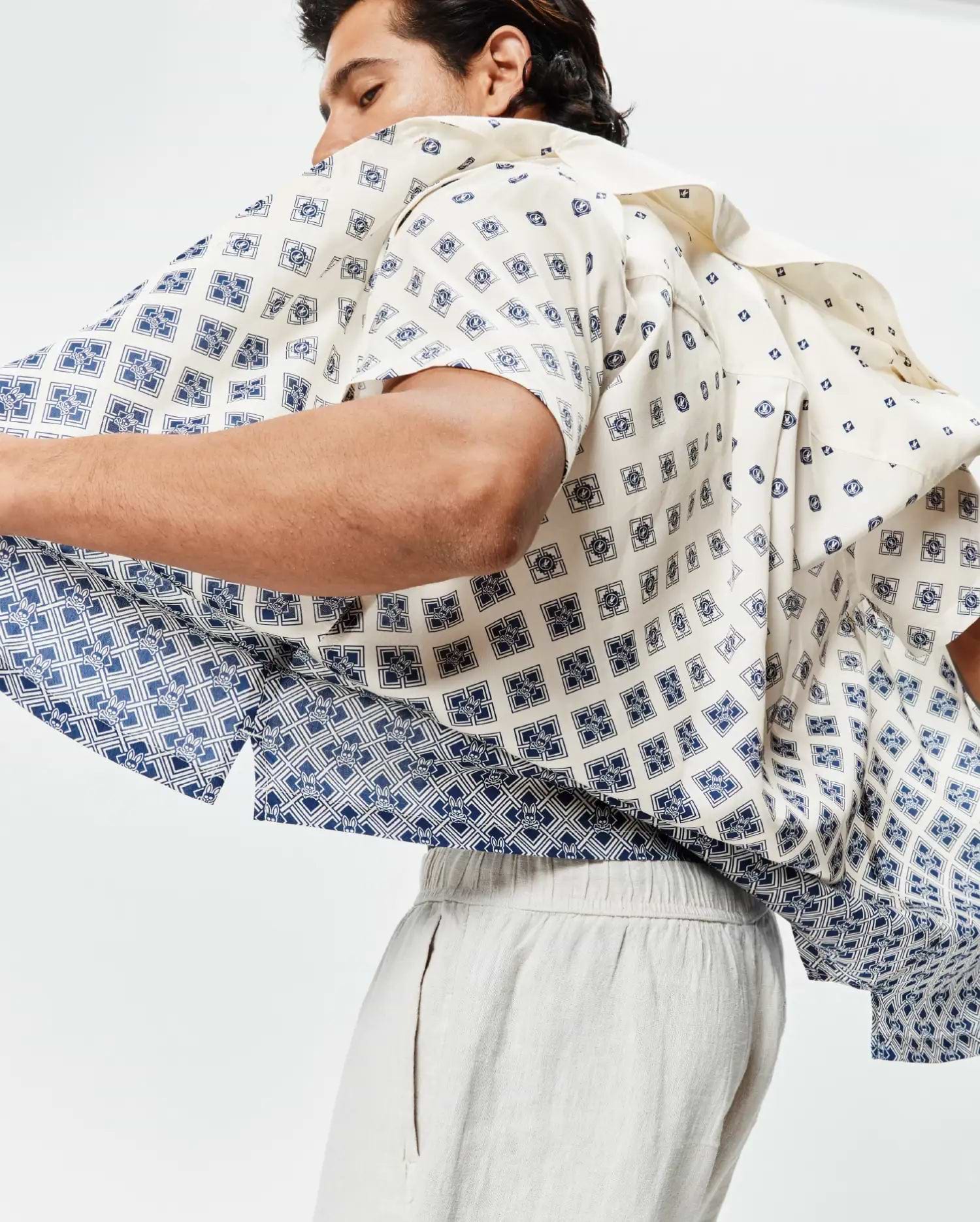 Mens Tobias Tencel Shirt, cream short-sleeve with a relaxed fit, featuring an all-over navy blue geometric pattern subtly incorporating small Psycho Bunny skull logos, transitioning to a denser pattern at the hem.