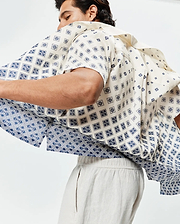 Mens Tobias Tencel Shirt, cream short-sleeve with a relaxed fit, featuring an all-over navy blue geometric pattern subtly incorporating small Psycho Bunny skull logos, transitioning to a denser pattern at the hem.