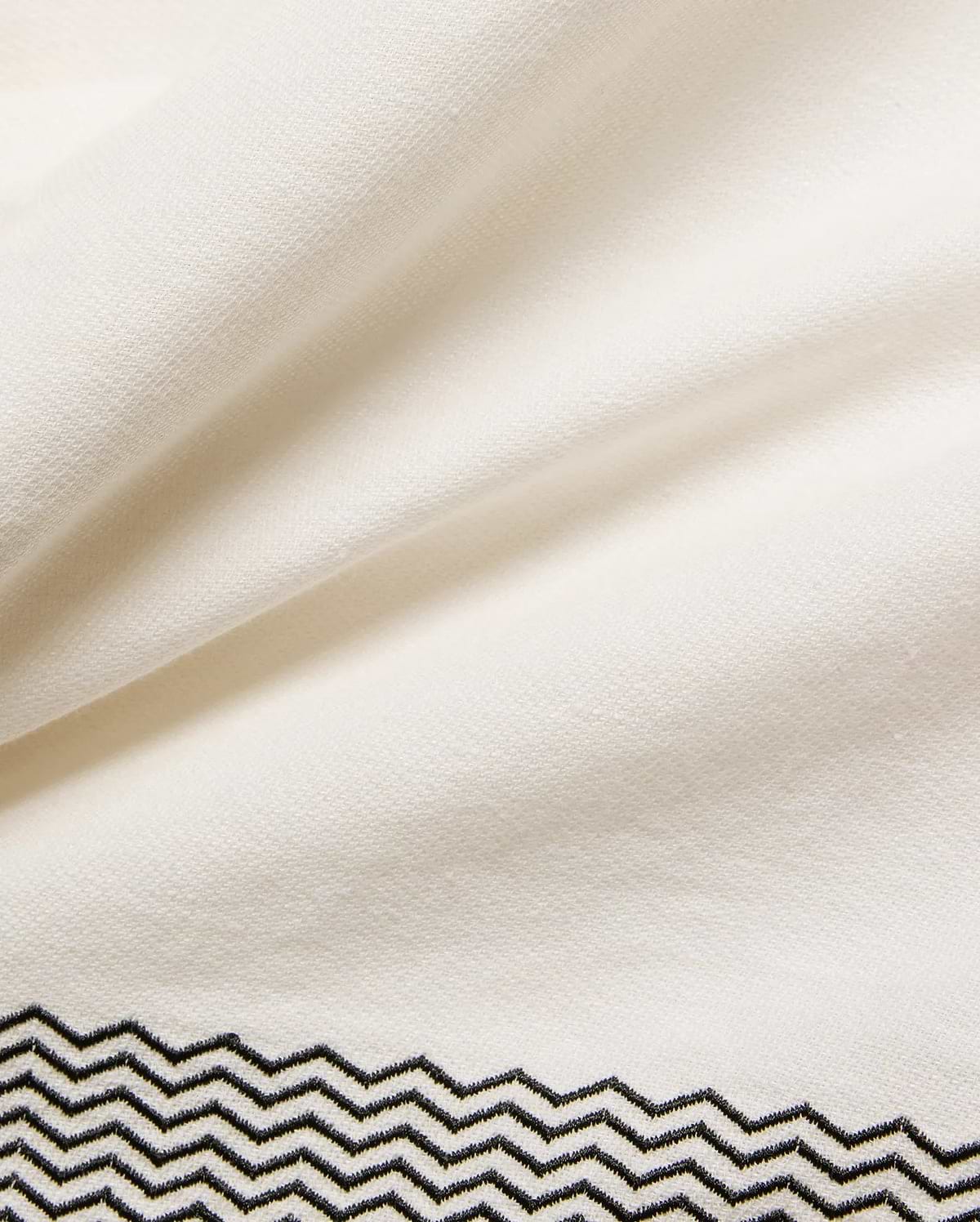 Mens Sunny Linen Shirt in crisp cream linen fabric, showcasing a refined black embroidered zigzag pattern with intricate stitching detail along the bottom hem. (6)