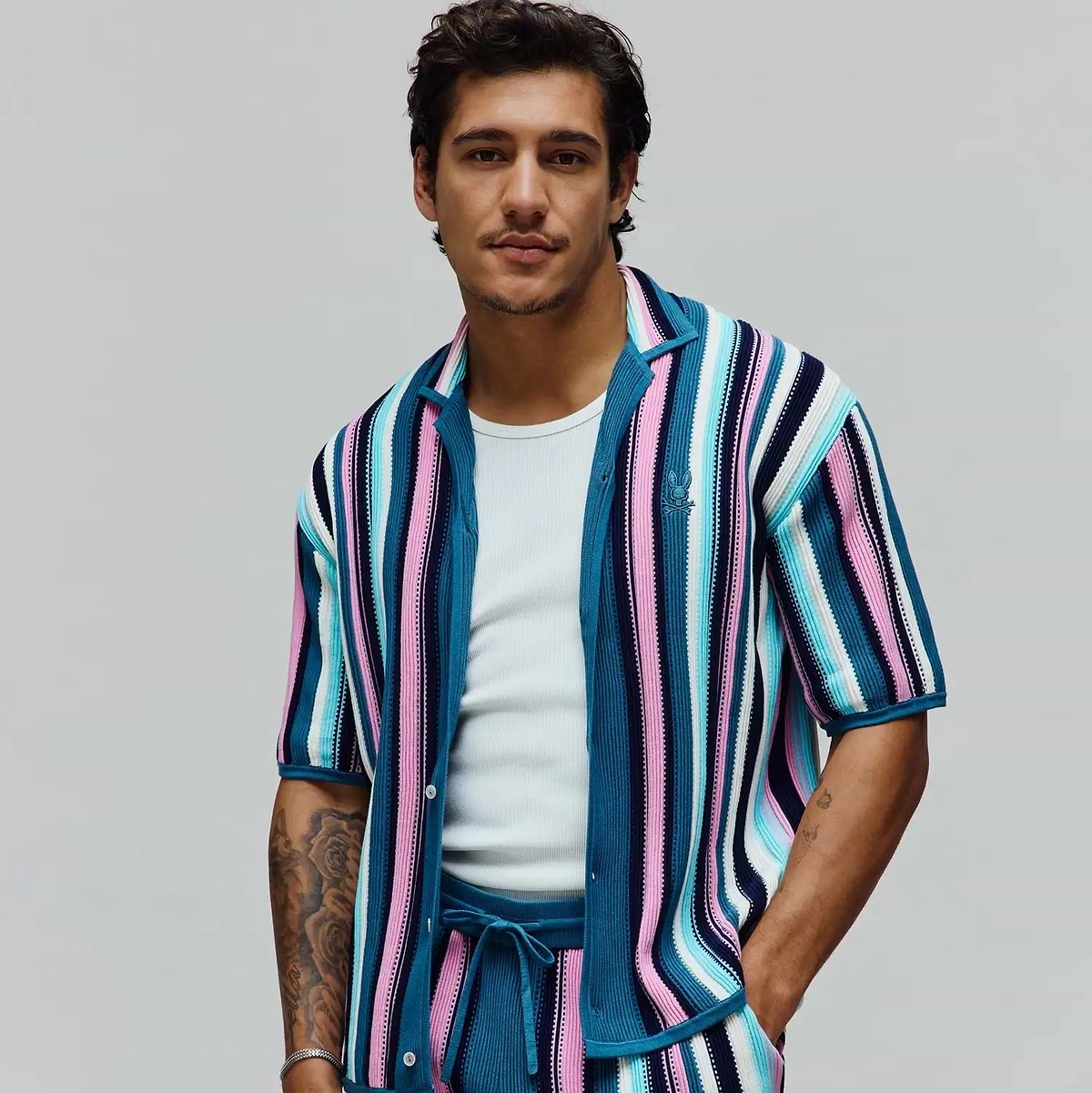 Mens Cliff Shirt, a vibrant knit short-sleeve shirt with teal, pink, light blue, white, and navy vertical stripes, featuring a small embroidered Psycho Bunny skull logo on the left chest. (1)