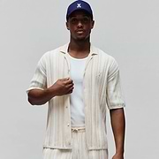 Mens Cliff Shirt, off-white relaxed fit short-sleeve button-up with beige vertical stripes, ribbed knit texture, and small embroidered Psycho Bunny skull logo on left chest. Matching shorts and navy cap with white bunny skull.