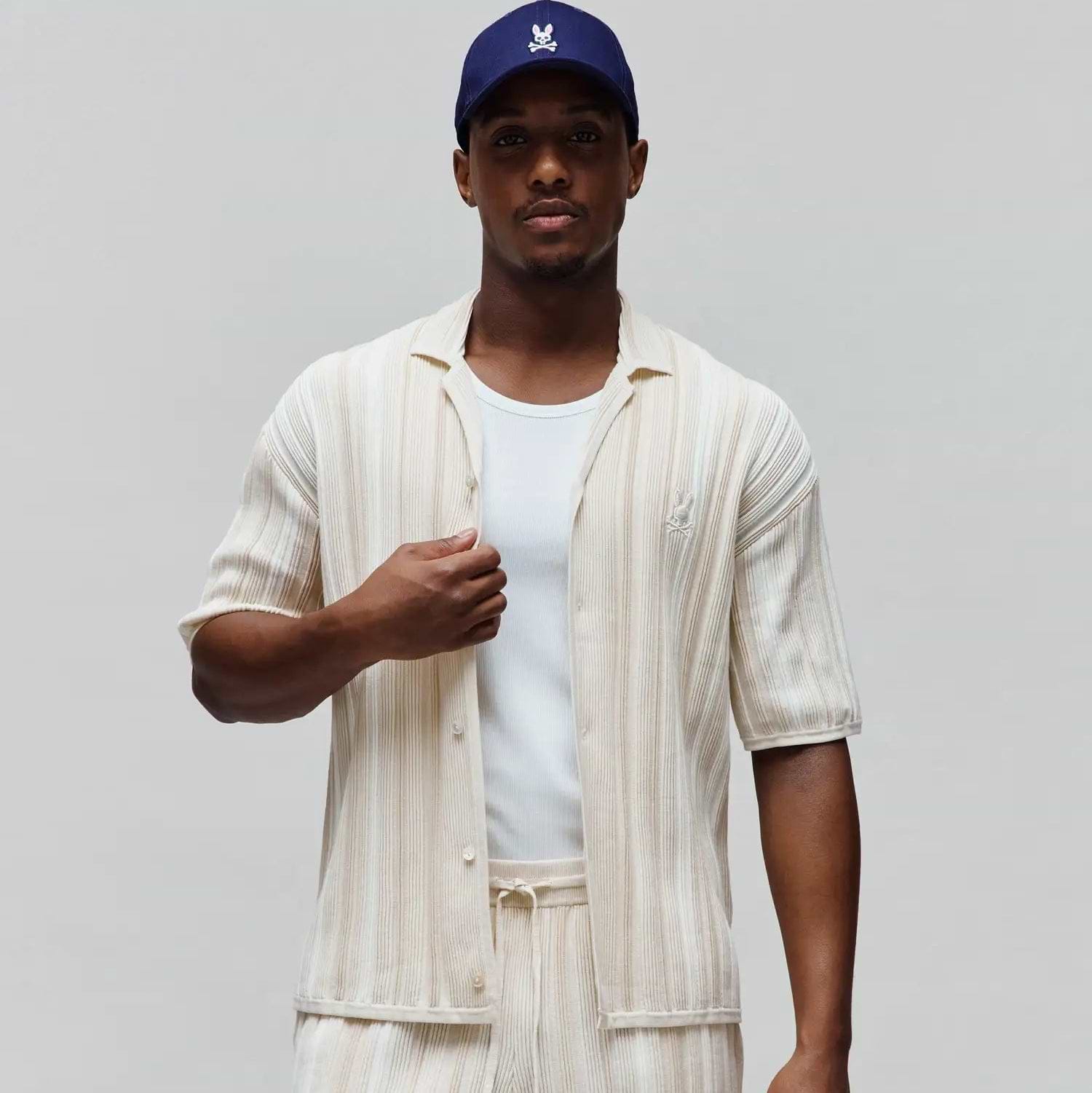 Mens Cliff Shirt, off-white relaxed fit short-sleeve button-up with beige vertical stripes, ribbed knit texture, and small embroidered Psycho Bunny skull logo on left chest. Matching shorts and navy cap with white bunny skull.