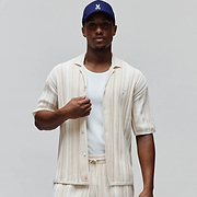 Mens Cliff Shirt, off-white relaxed fit short-sleeve button-up with beige vertical stripes, ribbed knit texture, and small embroidered Psycho Bunny skull logo on left chest. Matching shorts and navy cap with white bunny skull.