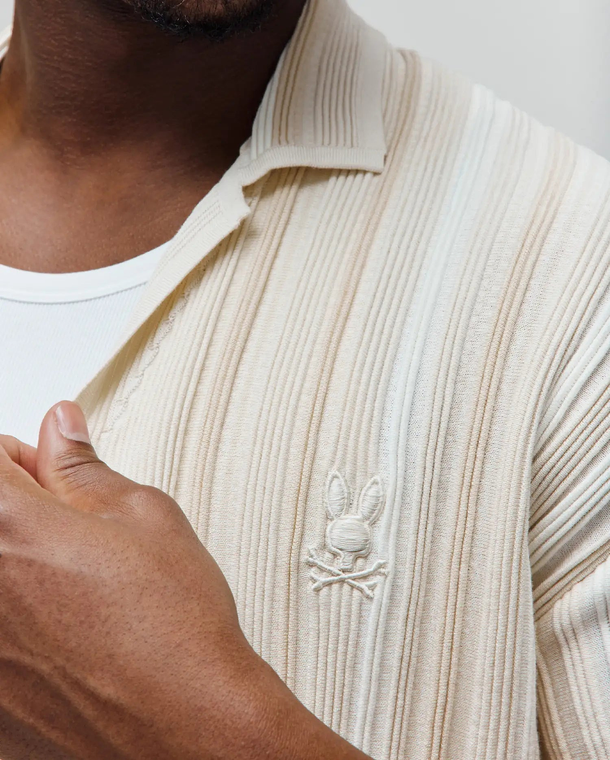 Mens Cliff Shirt, a cream ribbed knit polo with a classic fit. Features a subtle vertical texture and a small, tone-on-tone embroidered Psycho Bunny skull logo on the left chest.
