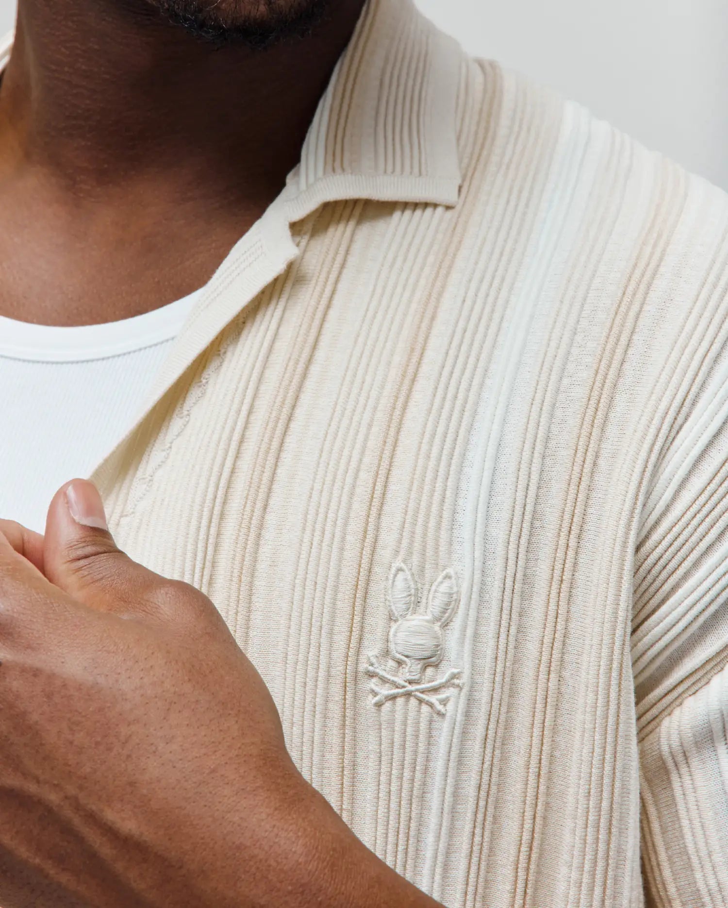 Mens Cliff Shirt, a cream ribbed knit polo with a classic fit. Features a subtle vertical texture and a small, tone-on-tone embroidered Psycho Bunny skull logo on the left chest.