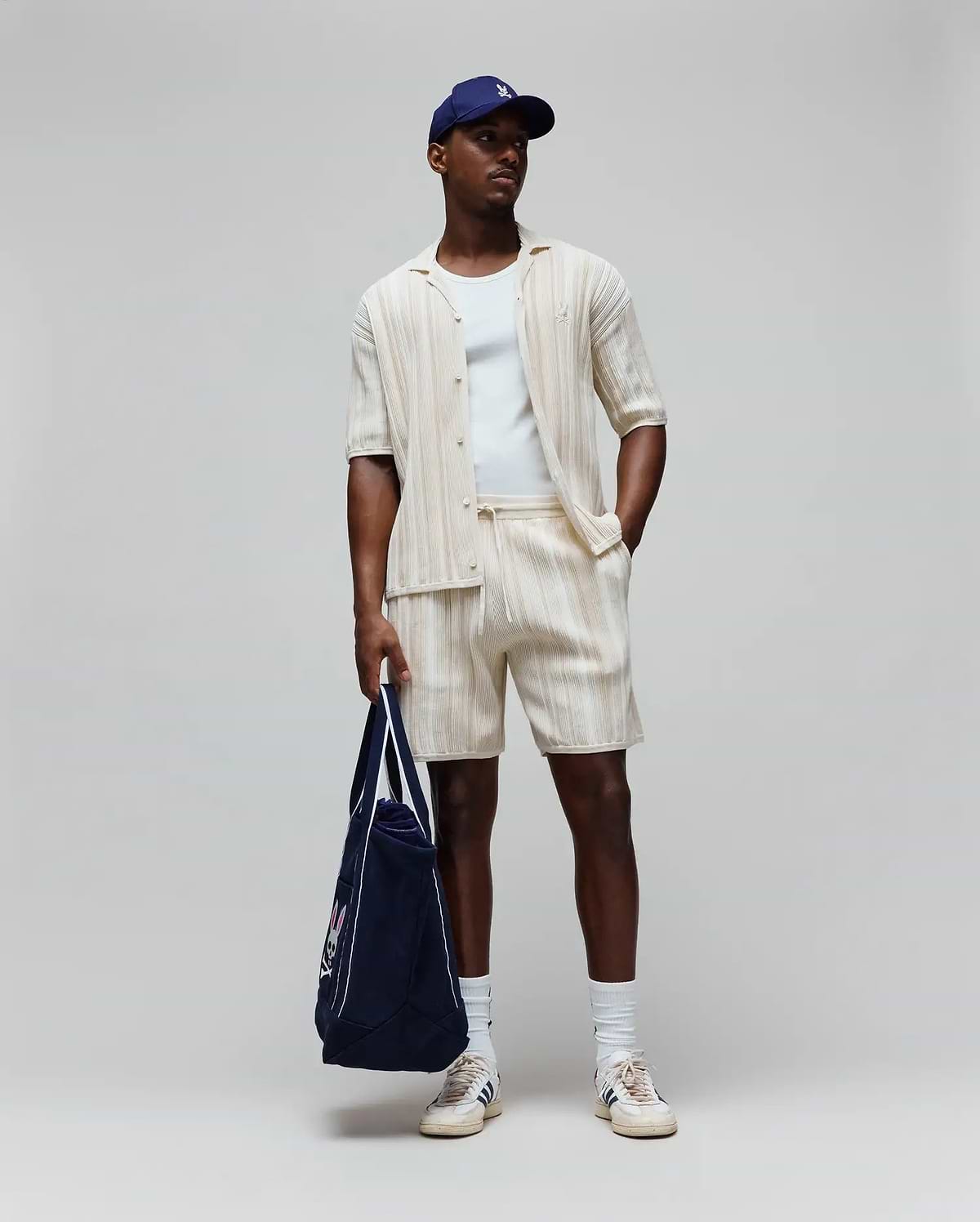 Mens Cliff Shirt, a cream ribbed shirt with small embroidered Psycho Bunny logo, matching relaxed drawstring shorts, white tee, navy cap with white logo, and navy tote with large Psycho Bunny graphic.
