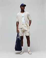 Mens Cliff Shirt, a cream ribbed shirt with small embroidered Psycho Bunny logo, matching relaxed drawstring shorts, white tee, navy cap with white logo, and navy tote with large Psycho Bunny graphic.