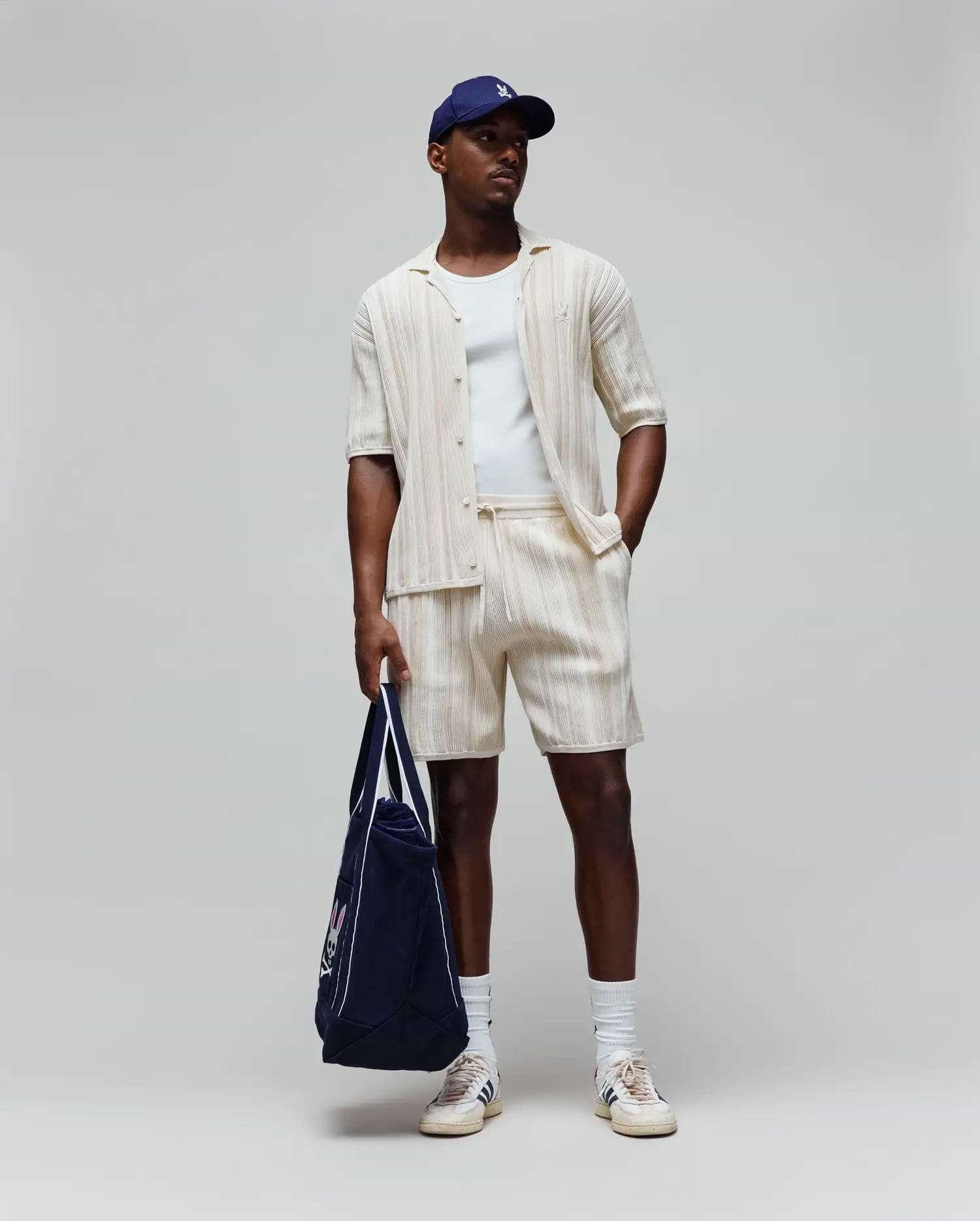 Mens Cliff Shirt, a cream ribbed shirt with small embroidered Psycho Bunny logo, matching relaxed drawstring shorts, white tee, navy cap with white logo, and navy tote with large Psycho Bunny graphic.