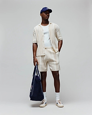 Mens Cliff Shirt, a cream ribbed shirt with small embroidered Psycho Bunny logo, matching relaxed drawstring shorts, white tee, navy cap with white logo, and navy tote with large Psycho Bunny graphic.
