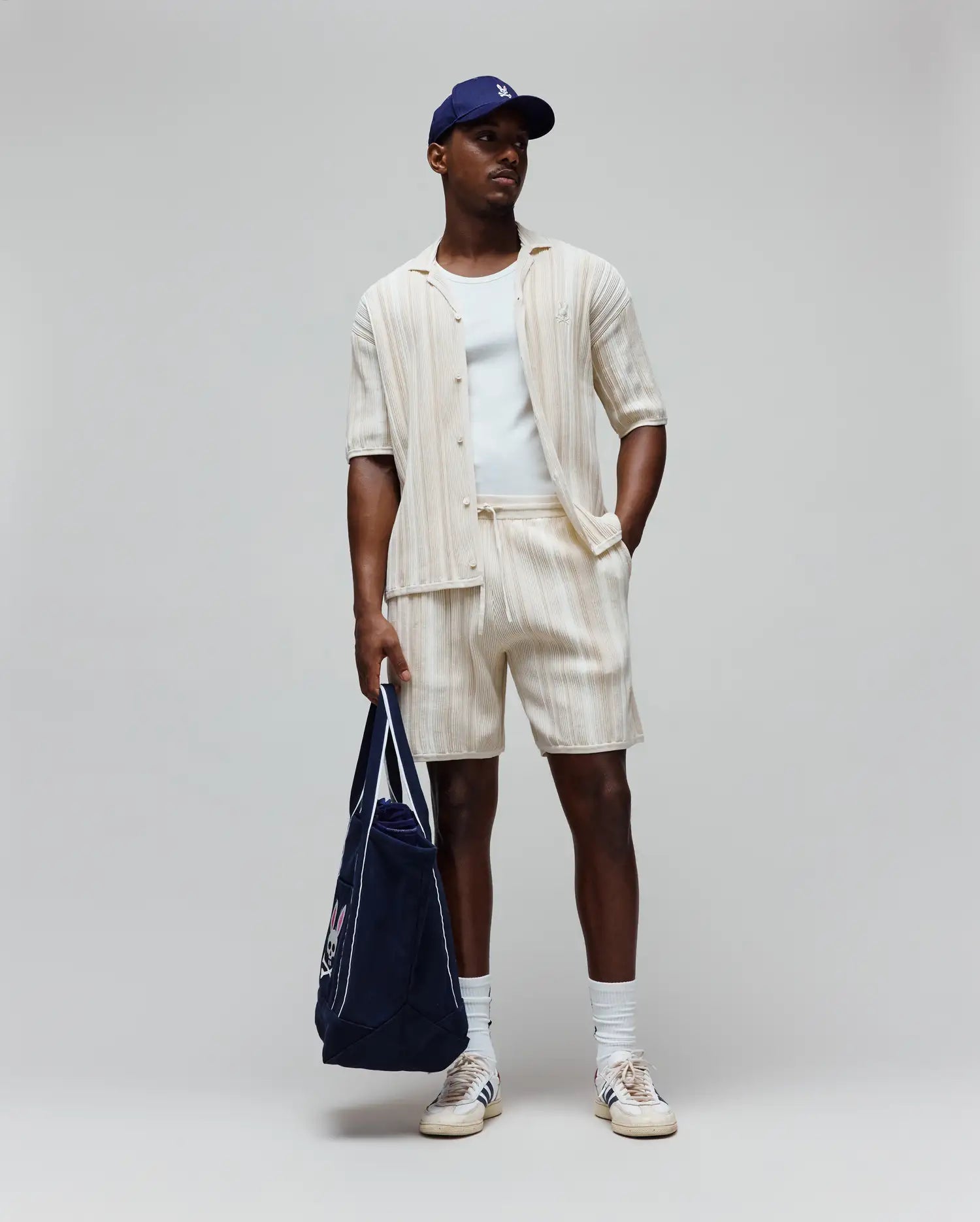 Mens Cliff Shirt, a cream ribbed shirt with small embroidered Psycho Bunny logo, matching relaxed drawstring shorts, white tee, navy cap with white logo, and navy tote with large Psycho Bunny graphic.