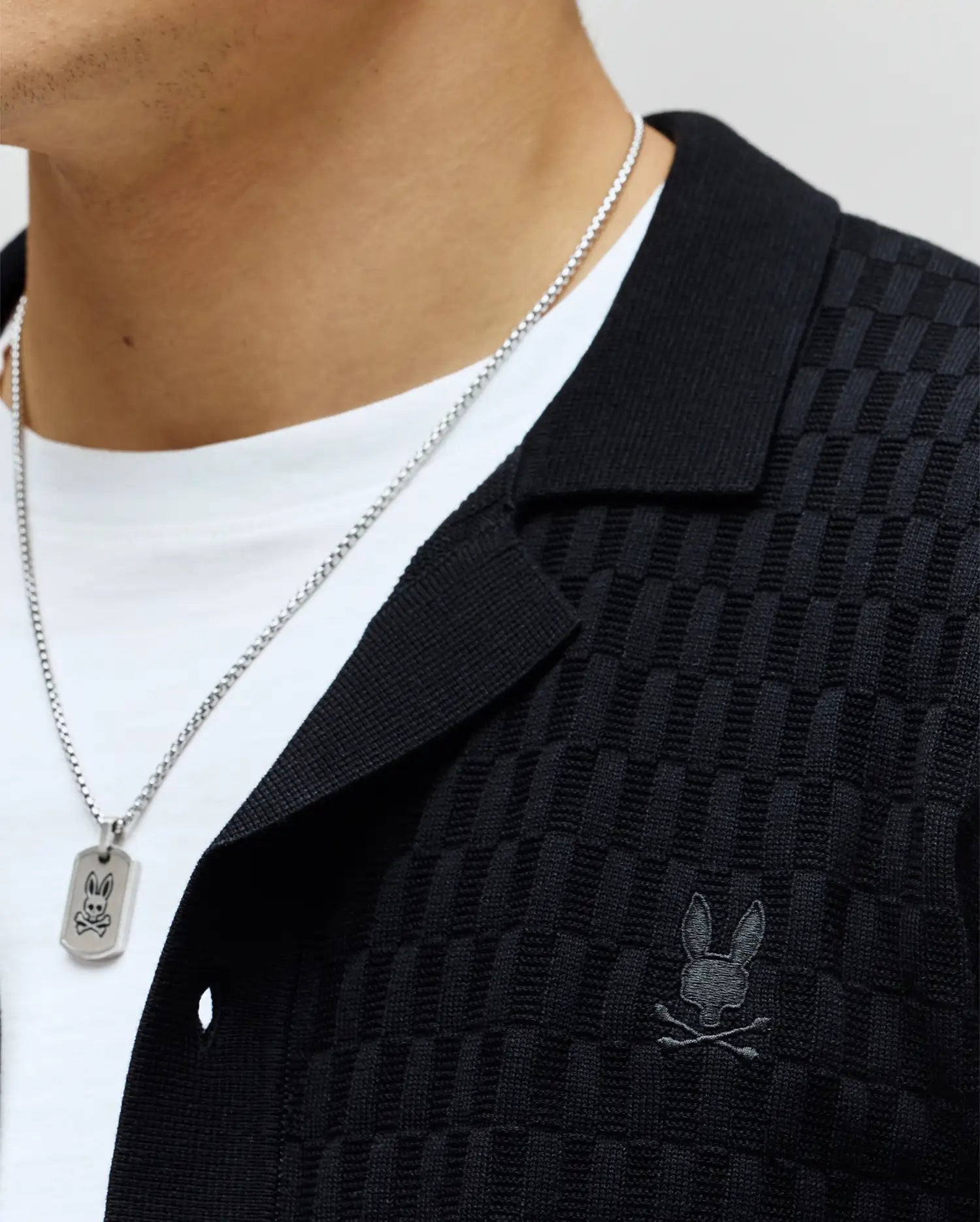 Mens Xandros Knit Shirt, black, featuring a refined checkerboard knit texture, classic collar, and a small tonal embroidered Psycho Bunny skull logo on the left chest.