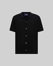 Mens Xandros Knit Shirt, black short-sleeve with a subtle geometric knit pattern, spread collar, white buttons, and a small embroidered black Psycho Bunny skull logo on the chest.