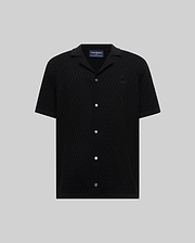 Mens Xandros Knit Shirt, black short-sleeve with a subtle geometric knit pattern, spread collar, white buttons, and a small embroidered black Psycho Bunny skull logo on the chest.