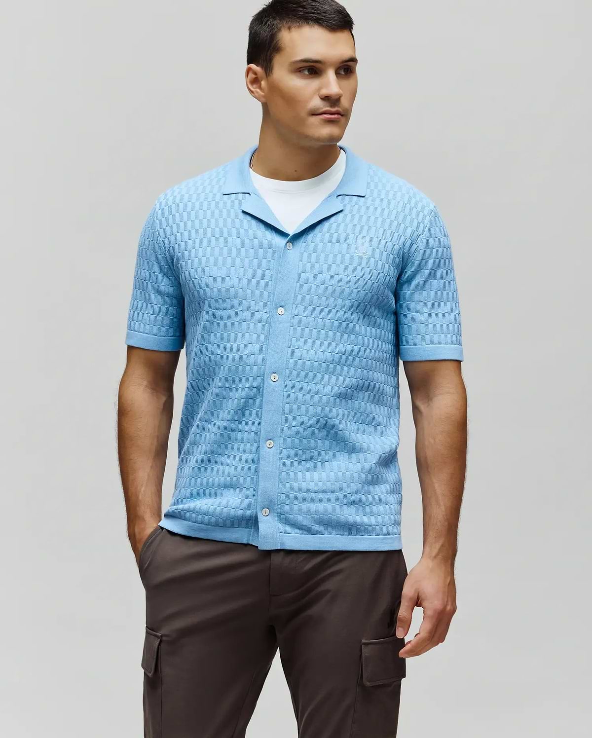 Mens Xandros Knit Shirt, a light blue classic fit short-sleeve knit shirt featuring a textured checkerboard pattern, full button placket with white buttons, spread collar, and a small embroidered Psycho Bunny skull logo on the left chest.