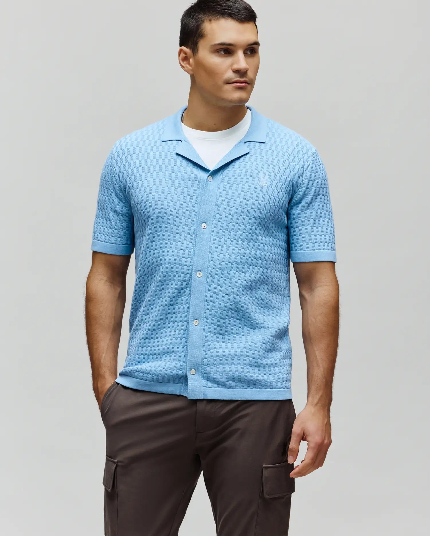 Mens Xandros Knit Shirt, a light blue classic fit short-sleeve knit shirt featuring a textured checkerboard pattern, full button placket with white buttons, spread collar, and a small embroidered Psycho Bunny skull logo on the left chest.