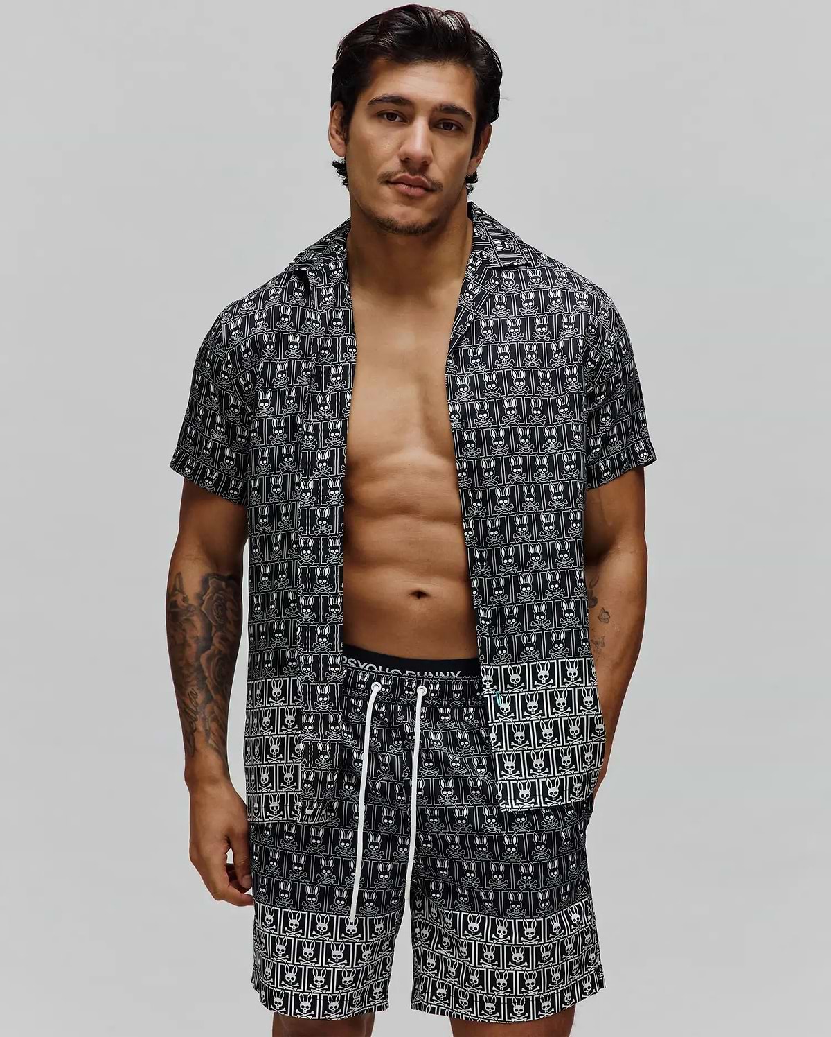 Mens Keon Tencel Shirt, a relaxed fit black short-sleeve button-up with an all-over white Psycho Bunny skull logo pattern, paired with matching shorts featuring white drawstrings. (1)