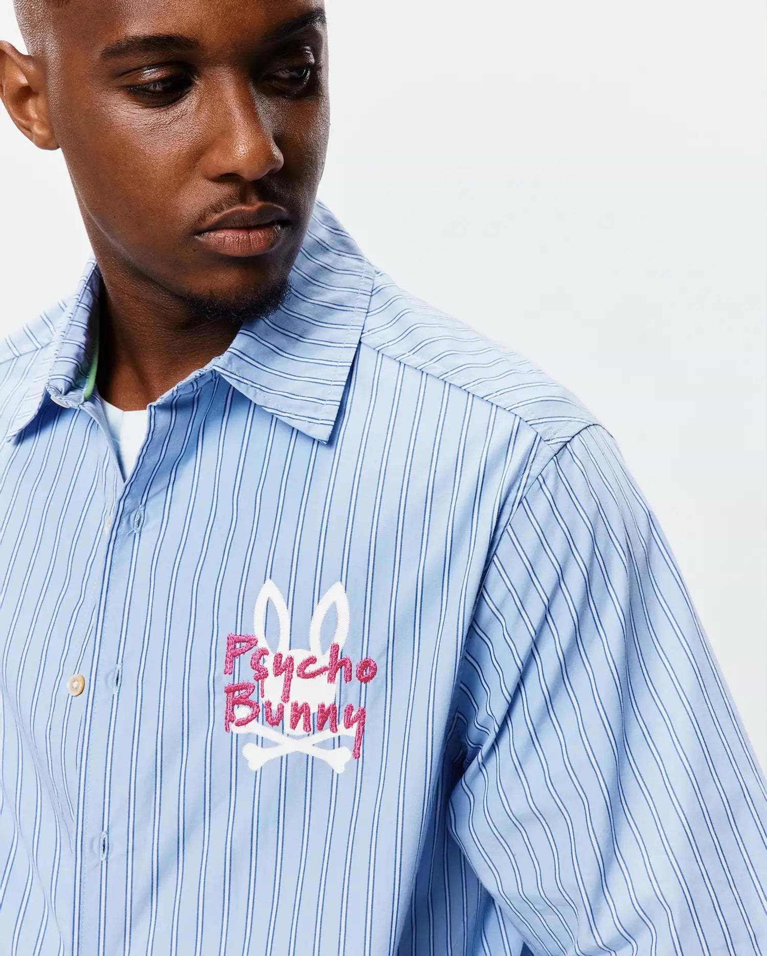 Mens Duncan Stripe Shirt, a light blue button-up with crisp dark blue and white vertical stripes. Features a large, embroidered Psycho Bunny skull logo in pink and white on the left chest, classic fit. (3)