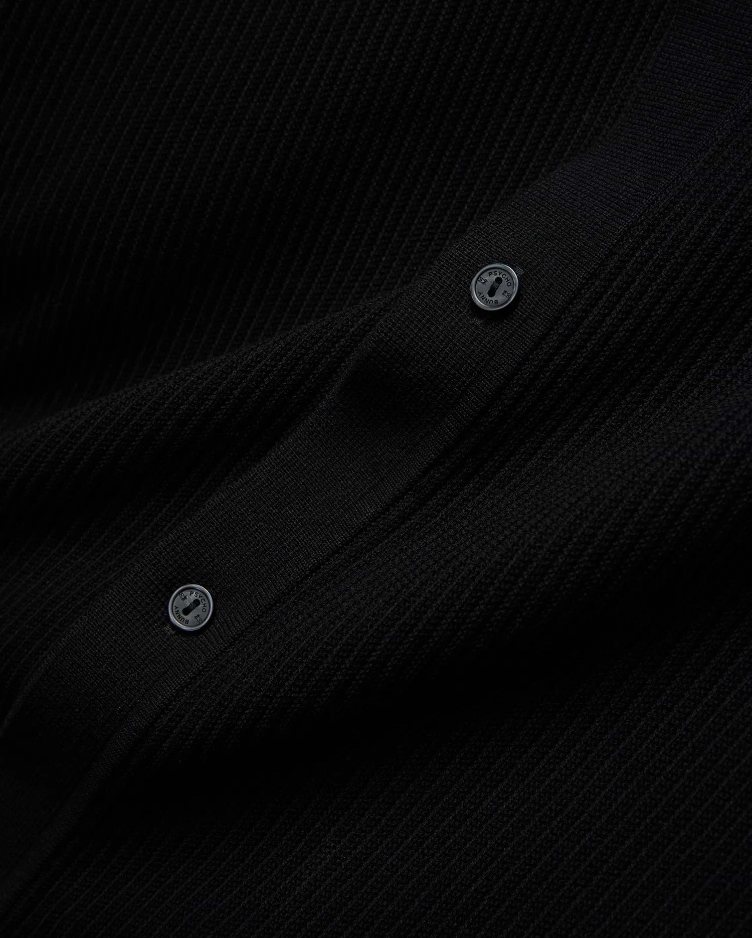 Mens Yann Knit Shirt, a sleek black premium ribbed knit garment, detailed with two dark buttons featuring the Psycho Bunny brand name embossed in a lighter tone.