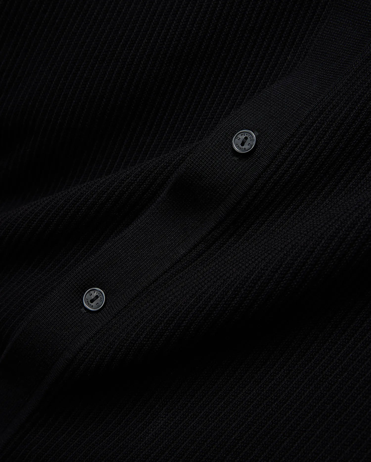 Mens Yann Knit Shirt, a sleek black premium ribbed knit garment, detailed with two dark buttons featuring the Psycho Bunny brand name embossed in a lighter tone.