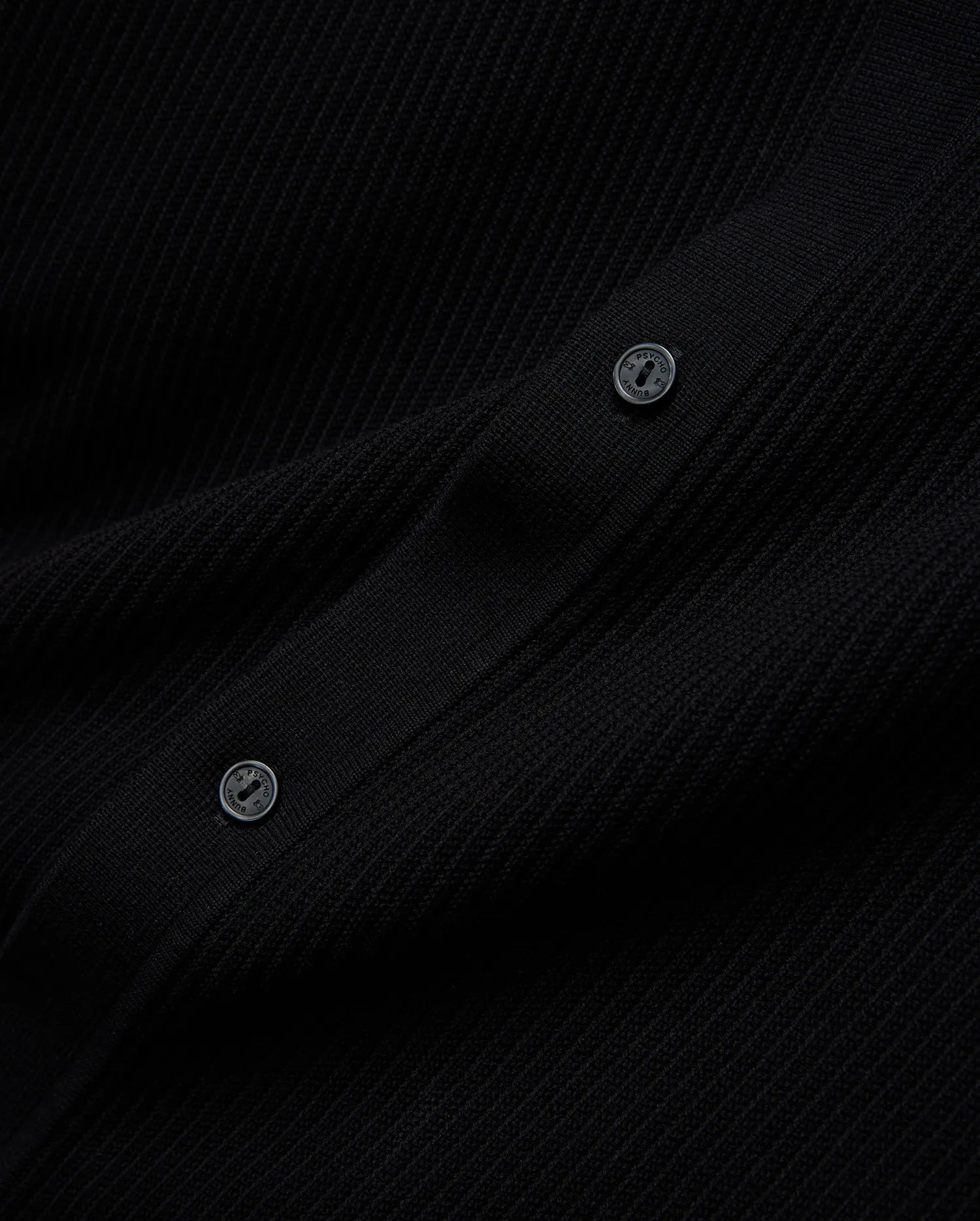 Mens Yann Knit Shirt, a sleek black premium ribbed knit garment, detailed with two dark buttons featuring the Psycho Bunny brand name embossed in a lighter tone.