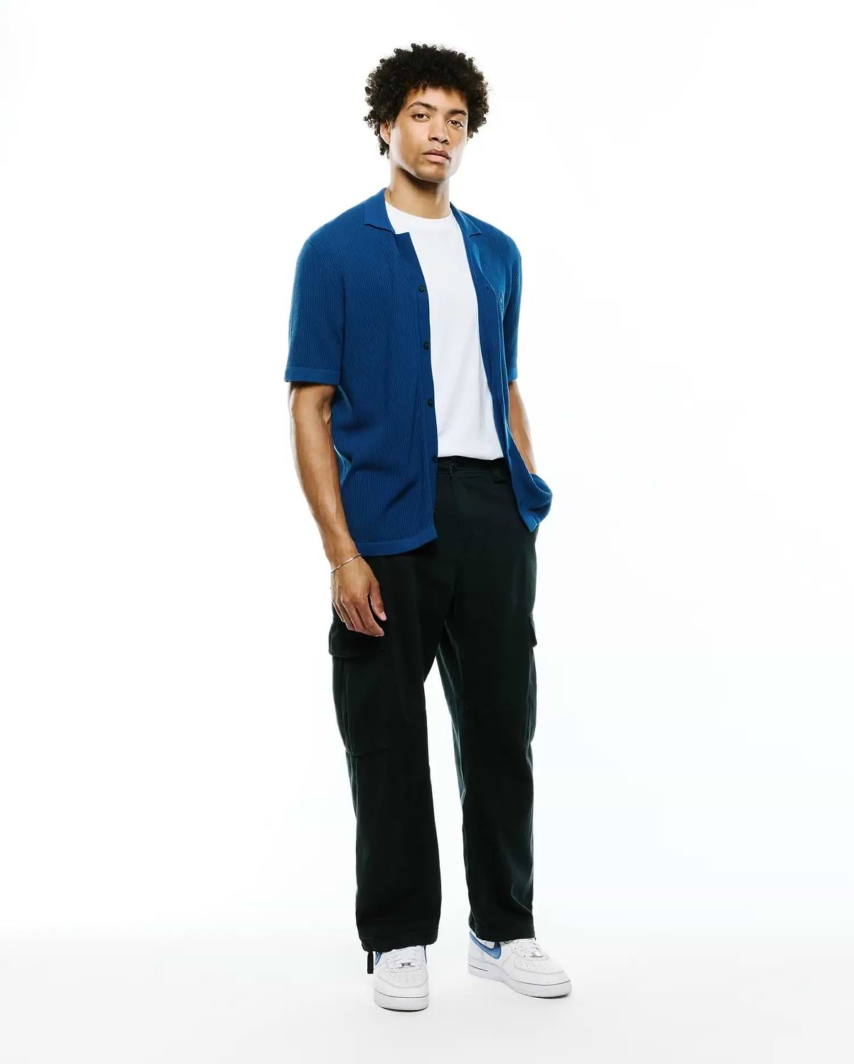 Mens Yann Knit Shirt: Premium vibrant blue ribbed knit short-sleeve button-up, relaxed fit, worn open over a white tee, with black cargo pants and white sneakers.