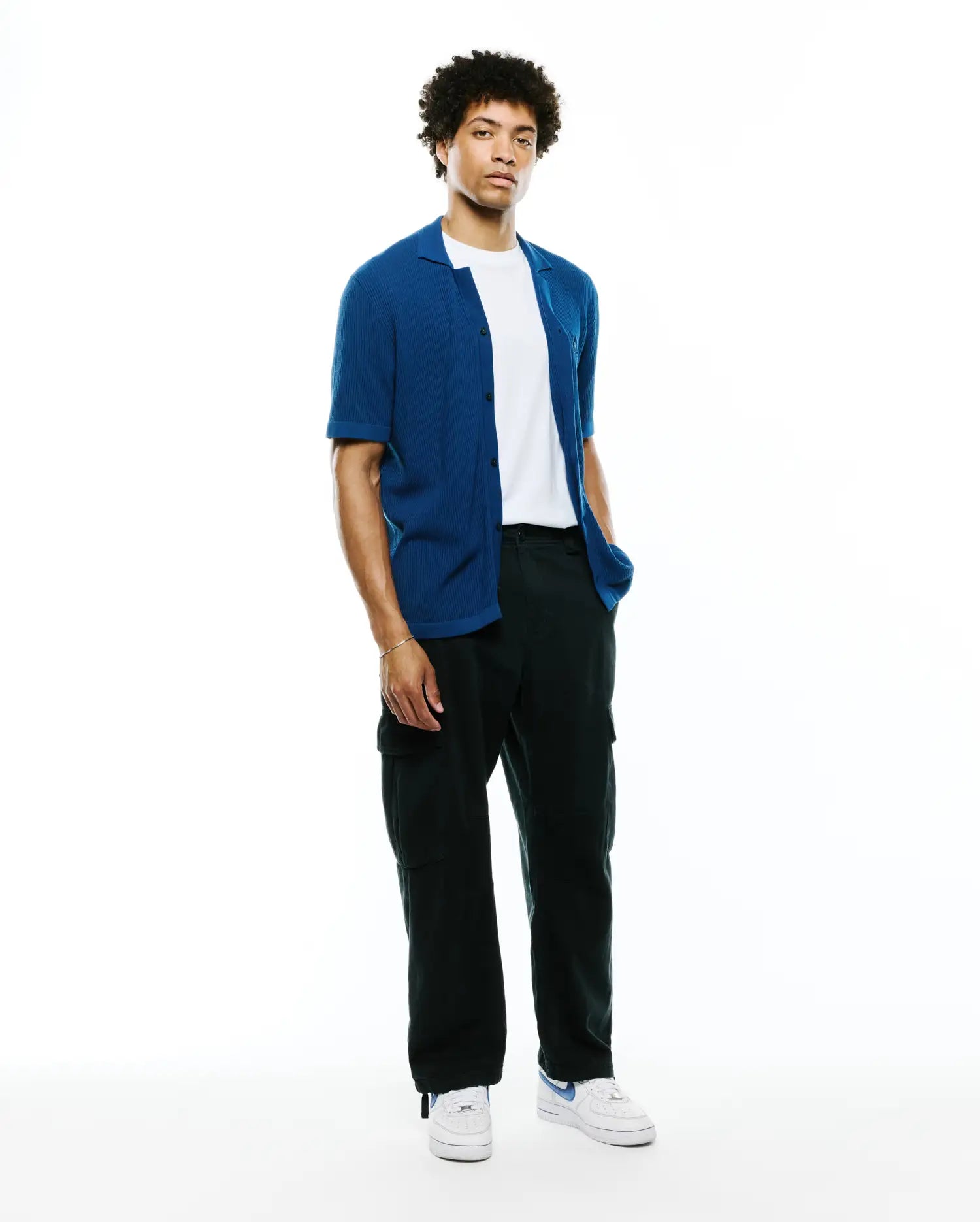 Mens Yann Knit Shirt: Premium vibrant blue ribbed knit short-sleeve button-up, relaxed fit, worn open over a white tee, with black cargo pants and white sneakers.