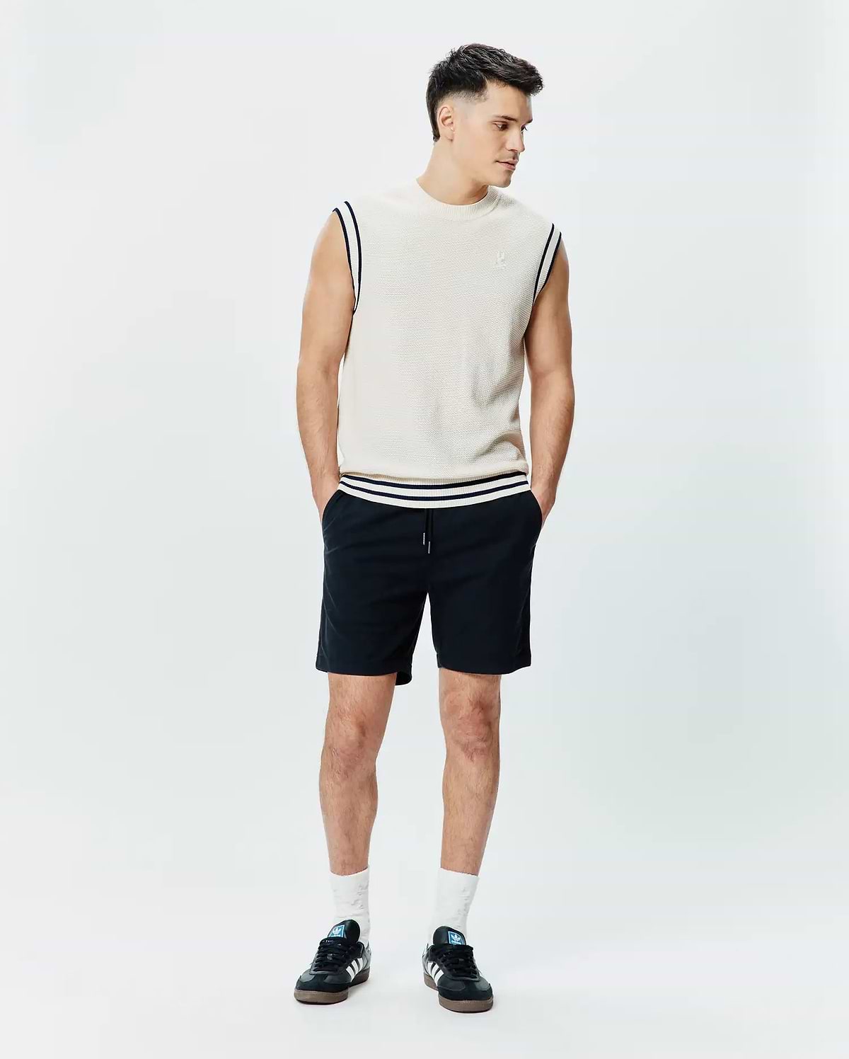 Mens Peter Knit Vest in cream, featuring a classic fit, fine knit texture, navy and white striped armhole and hem trim, and a small embroidered Psycho Bunny skull on the left chest, paired with black drawstring shorts.