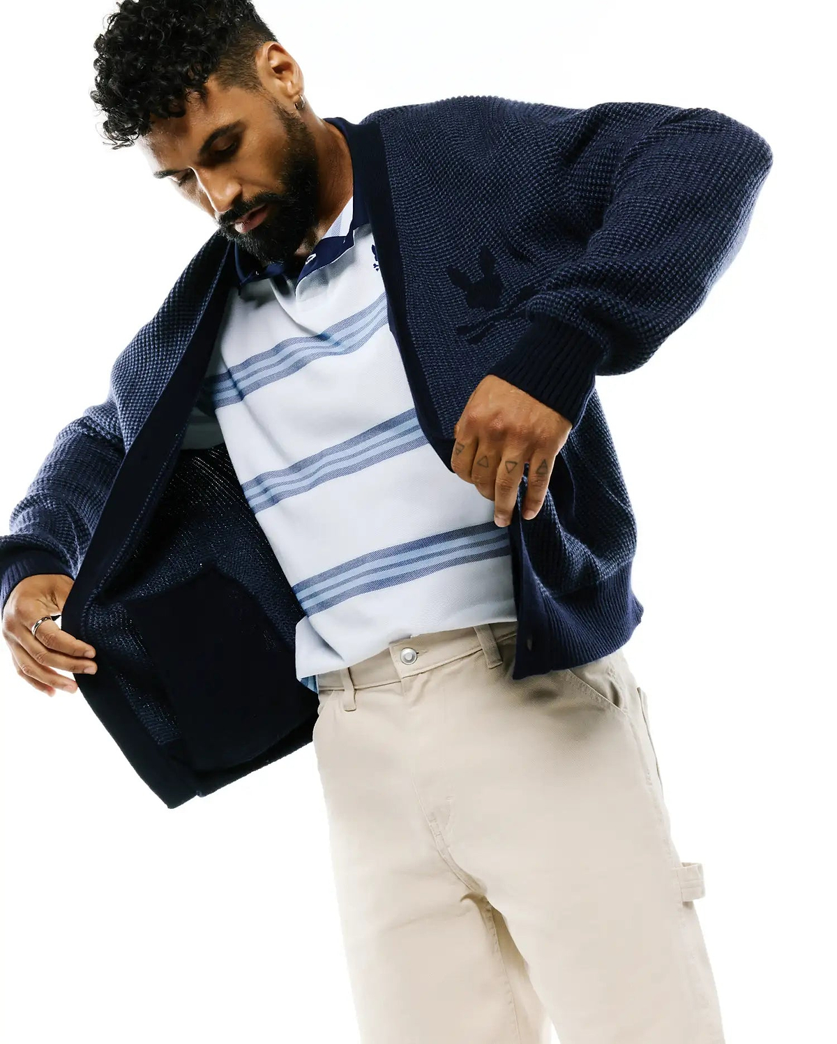 Mens Atkinson Cardigan, a navy waffle knit cardigan with a large tonal Psycho Bunny skull logo on the left chest, layered over a white and blue striped pique polo and beige twill carpenter pants.