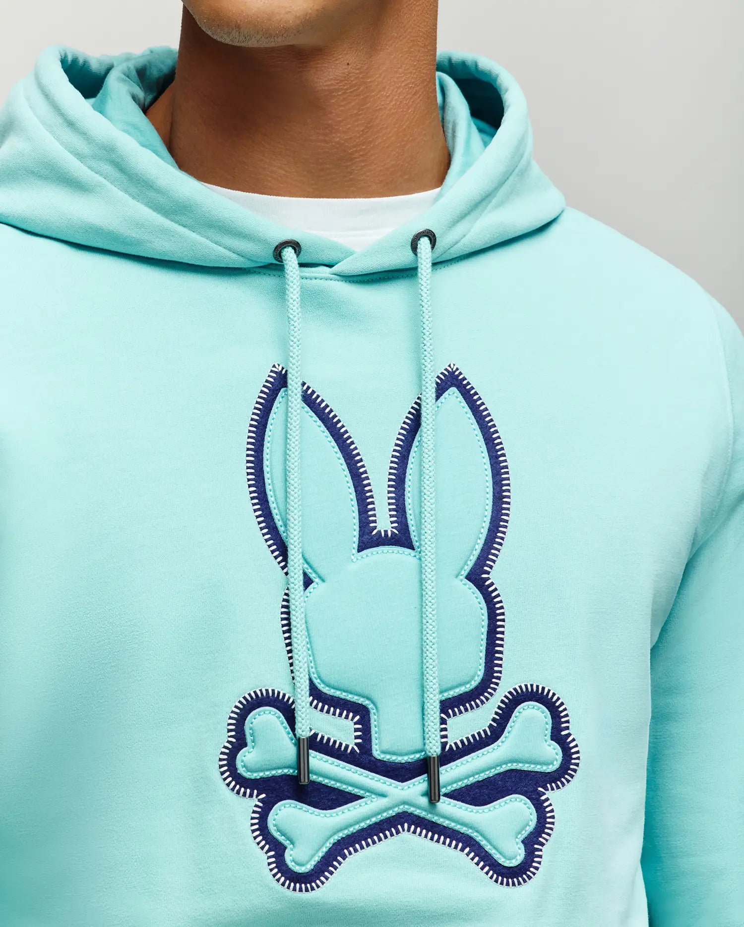 Mens Gage Patch Hoodie in aqua soft fleece, featuring a relaxed fit and a large navy-outlined bunny skull appliqué logo with white stitching on the chest, plus a matching drawstring hood.
