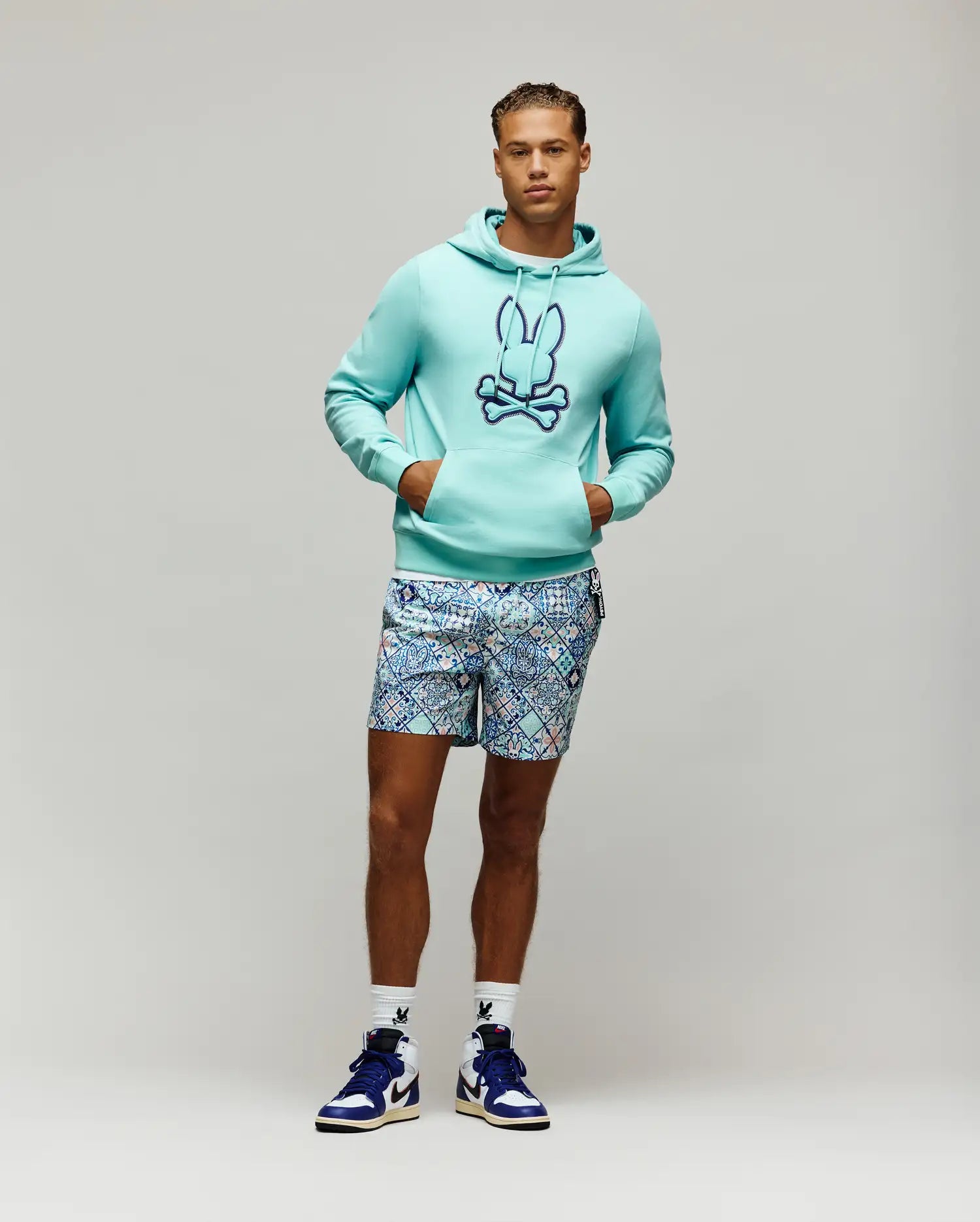 Aqua French terry Mens Gage Patch Hoodie, classic fit, with a large embroidered dark blue Psycho Bunny skull logo on the chest, paired with patterned blue and white shorts and white logo socks.