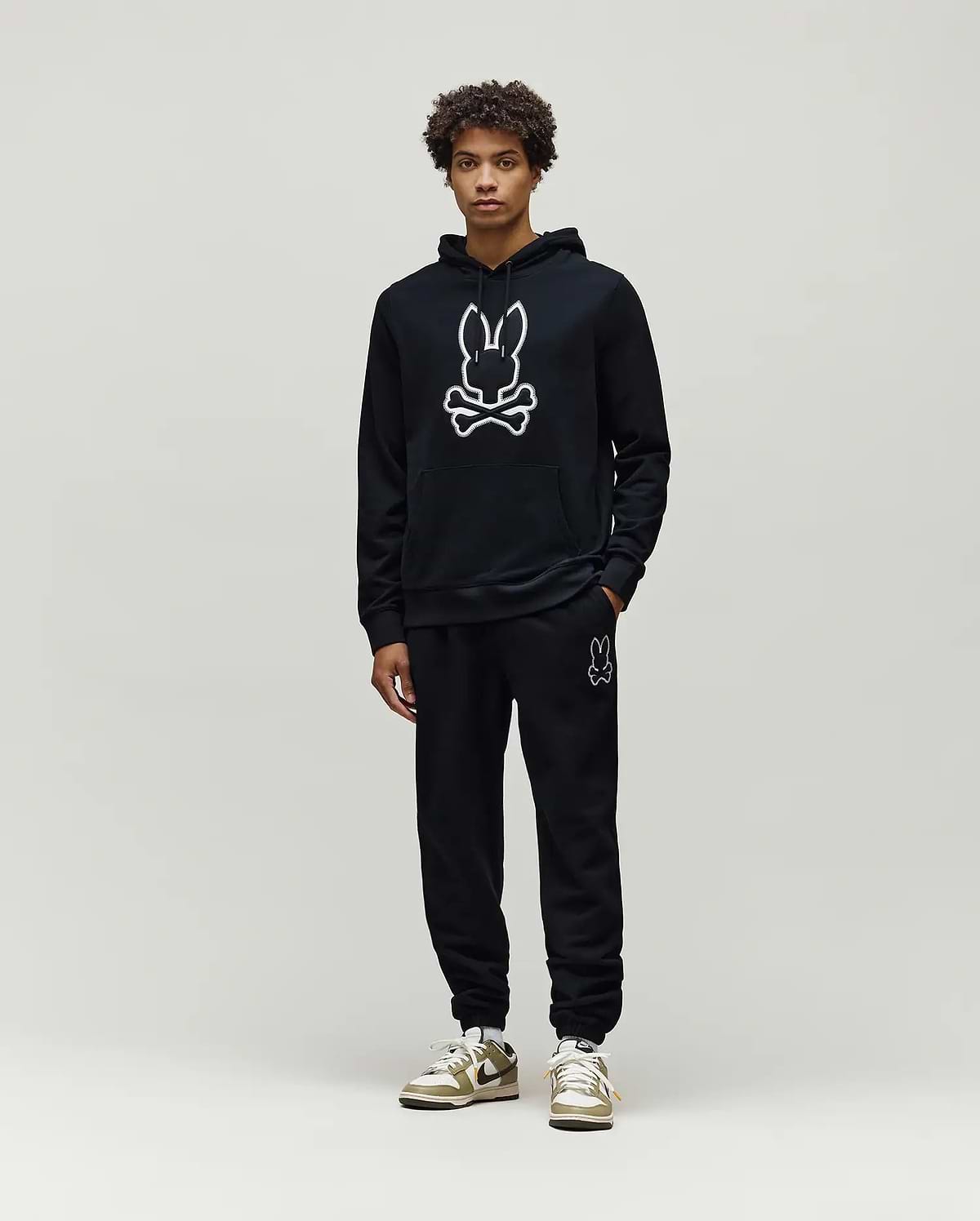Mens Gage Patch Hoodie: Black relaxed fit French terry hoodie with a large white outlined Psycho Bunny skull logo on the chest, kangaroo pocket, and ribbed cuffs, paired with matching black sweatpants.